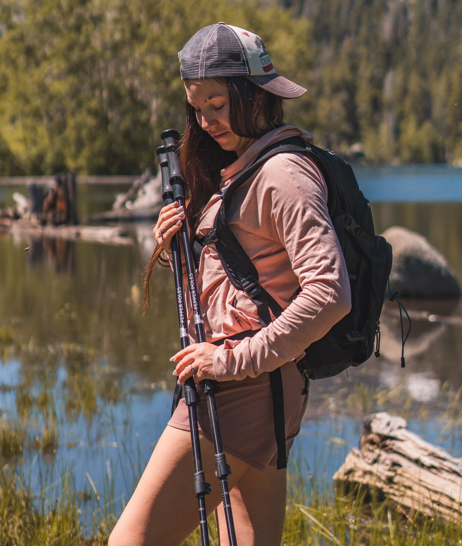 A woman hiker in a pink outfit and a backpack holds a pair of Pepper Poles near a scenic mountain lake, surrounded by lush greenery.