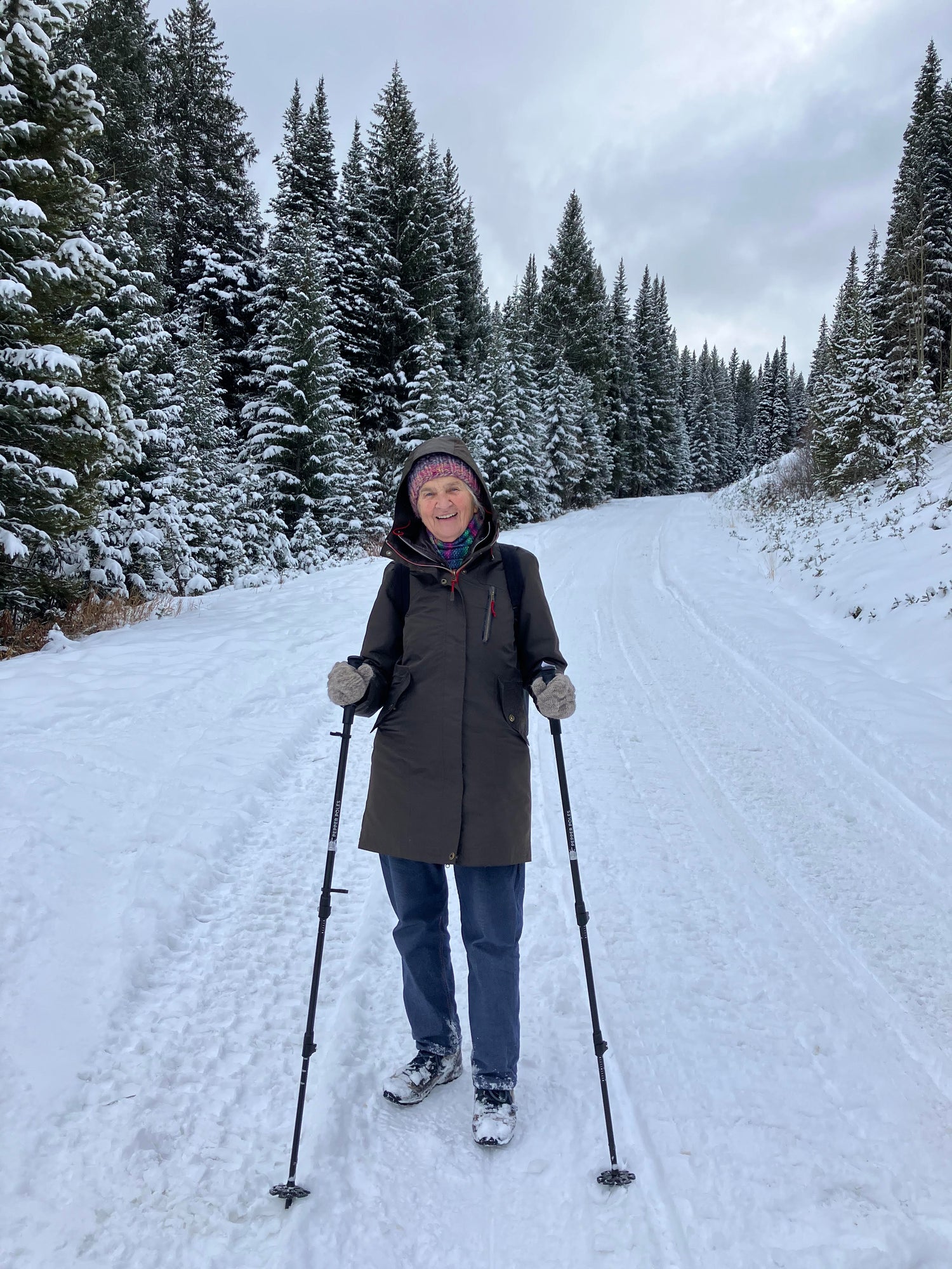 An elderly woman smiles while standing on a snow-covered trail surrounded by tall evergreen trees. She is dressed in a warm winter coat, a knitted hat, gloves, and jeans, and is using a pair of Pepper Poles for stability and safety. The snowy landscape and overcast sky create a serene winter hiking scene, emphasizing the versatility of Pepper Poles in various outdoor conditions.
