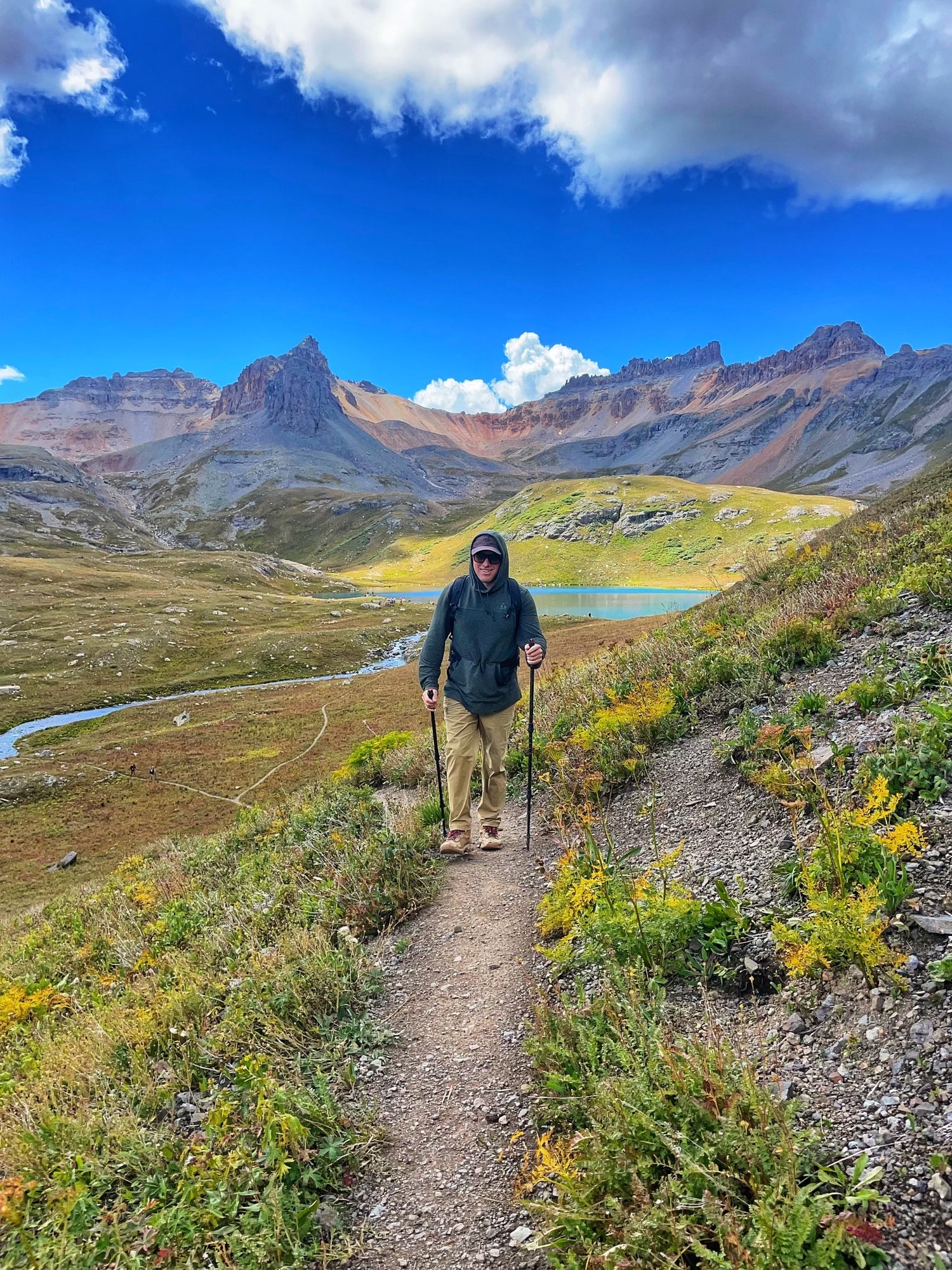 A hiker, dressed in a dark hoodie, sunglasses, and khaki pants, treks uphill on a narrow dirt trail using Pepper Poles, hiking poles equipped with discreet, built-in pepper spray for safety. The rugged alpine landscape features lush green vegetation, yellow wildflowers, and a winding trail leading toward a pristine mountain lake. Jagged peaks rise against a vivid blue sky with scattered clouds. The Pepper Poles provide both stability and security, ensuring the hiker is Ready for Anything.