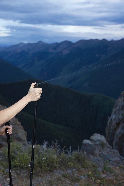 A hiker demonstrates the built-in pepper spray feature of Pepper Poles, spraying a fine mist into the air against a backdrop of rugged mountain peaks and dense forests. The hiker grips the pole firmly, with one hand holding the second pole for stability. The scene highlights the dual-purpose design of Pepper Poles, providing both hiking support and personal safety in remote outdoor environments.