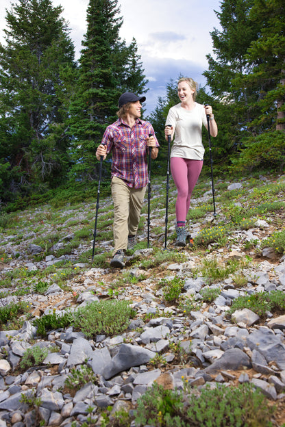 A man and a woman hike together on a rocky mountain trail, using Pepper Poles for stability and personal safety. They are smiling and engaged in conversation, enjoying their outdoor adventure. The man wears a plaid shirt, khaki pants, and a black cap, while the woman is dressed in a light-colored t-shirt and pink leggings. The lush evergreen trees and rugged terrain emphasize the versatility of Pepper Poles, designed to provide both hiking support and discreet self-defense with built-in pepper spray.